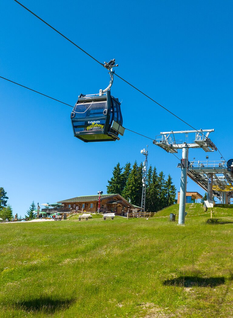 Two cable cars approach Rittisberg top station with hut, play area and green meadow under a clear blue sky. | © Rittis-Lift Engelhardt GmbH & Co KG