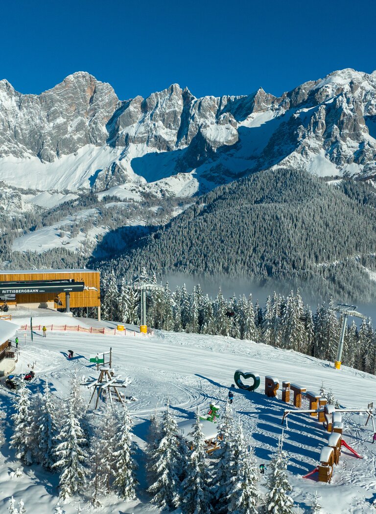 Rittisstadl and cable car station in snowy forest with playground and Dachstein mountains in winter sun. | © Rittis-Lift Engelhardt GmbH & Co KG