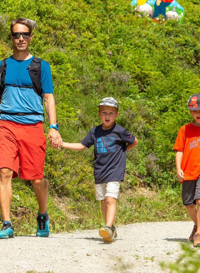 Family hikes a forest trail lined with colorful fairy tale figures like dwarfs on a sunny green hillside. | © Rittis-Lift Engelhardt GmbH & Co KG