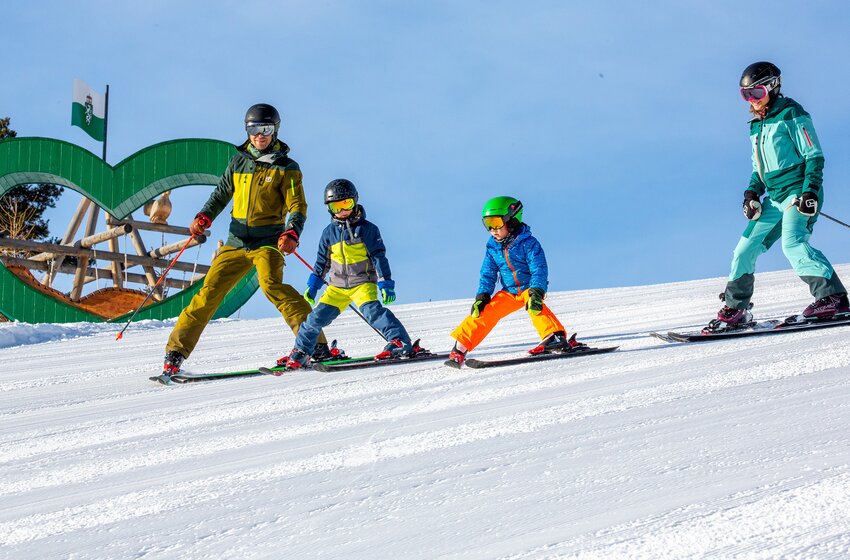 Family skis downhill in snowplow formation, with green wooden heart and playground at Rittisberg in the background. | © Rittis-Lift Engelhardt GmbH & Co KG