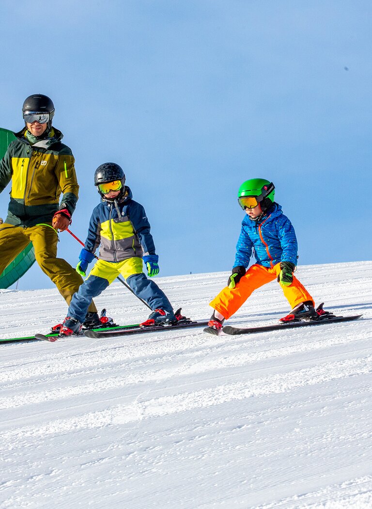 Family skis downhill in snowplow formation, with green wooden heart and playground at Rittisberg in the background. | © Rittis-Lift Engelhardt GmbH & Co KG