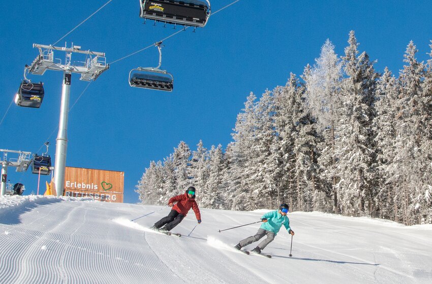 Two skiers skiing below the Rittisberg Erlebnisbahn lift | © Rittis-Lift Engelhardt GmbH & Co KG Two skiers enjoy a sunny descent below the Rittisberg Erlebnisbahn, with snowy forest beside the slope. | © Rittis-Lift Engelhardt GmbH & Co KG