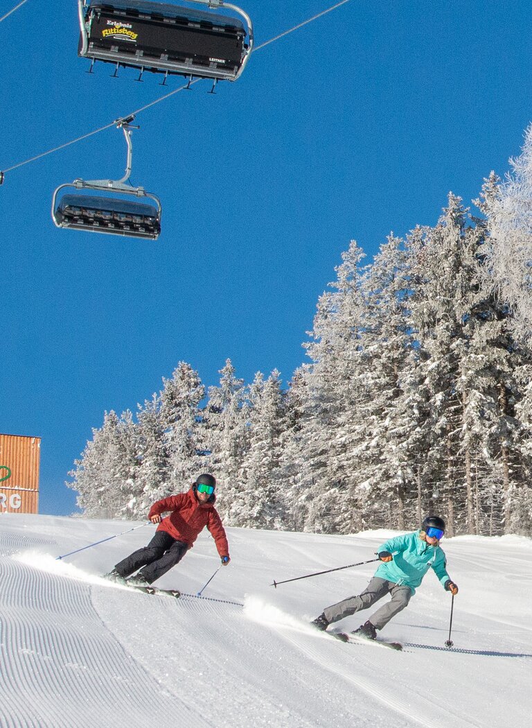 Two skiers enjoy a sunny descent below the Rittisberg Erlebnisbahn, with snowy forest beside the slope. | © Rittis-Lift Engelhardt GmbH & Co KG