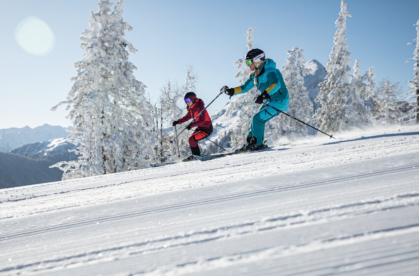 Two skiers descend side by side on a freshly groomed slope surrounded by snowy trees and mountain scenery. | © Reiteralm Bergbahnen GmbH & Co.KG