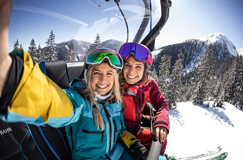 Two smiling skiers take a selfie on the chairlift with snowy trees and a sunny mountain backdrop. | © Reiteralm Bergbahnen GmbH & Co.KG