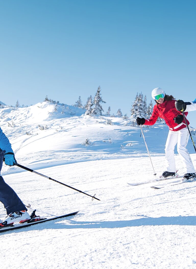 Skigruppe fährt gemeinsam eine sonnige Piste in Ski amadé hinab | © Ski amadé GmbH Drei Skifahrer fahren nebeneinander auf einer sonnigen, verschneiten Piste mit Sesselliften im Hintergrund in Ski amadé. | © Ski amadé GmbH