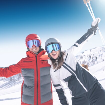 Two smiling skiers stand side by side, arms raised, wearing helmets and goggles against a sunny snowy mountain backdrop. | © Ski amadé GmbH