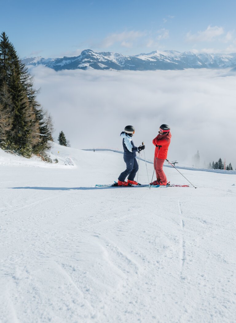 Zwei Skifahrer stehen auf breiter Piste über dichtem Nebelmeer, Berge ragen unter blauem Himmel hervor | © Ski amadé GmbH